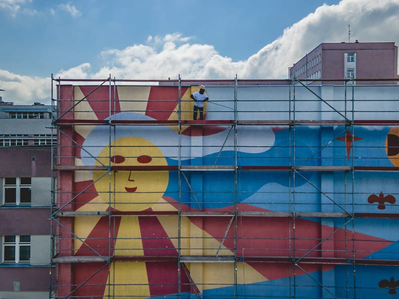 Dawny mural ze słońcem na rusztowaniu, w trakcie przemalowywania